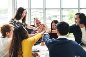 Coworkers gathered around a conference room table holding their hands together in the middle.